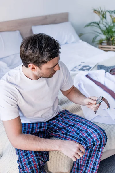 Красивий чоловік у піжамах тримає годинник, сидячи на постільній білизні вдома — стокове фото