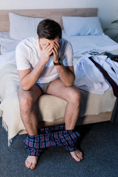 Schläfriger Mann mit den Händen im Gesicht sitzt auf Bettwäsche in der Nähe von Kleidung — Stockfoto