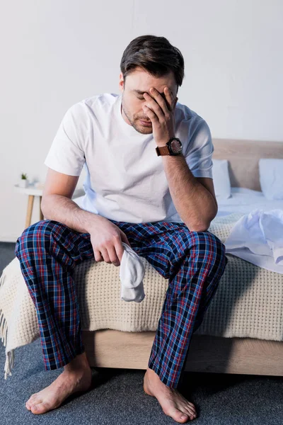 Schöner schläfriger Mann sitzt auf Bettwäsche und hält weiße Socken — Stockfoto