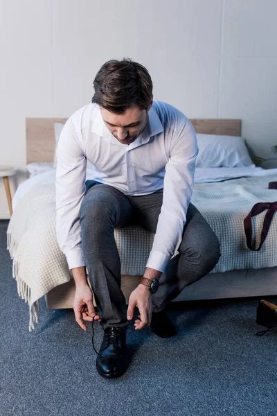 Schöner Mann zieht schwarze Schuhe an, während er zu Hause auf Bettwäsche sitzt — Stockfoto