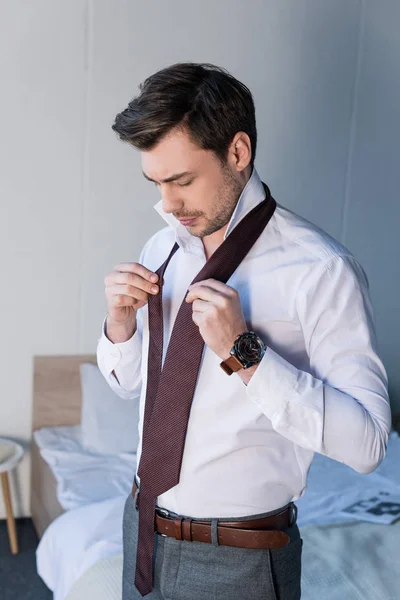 Schöner selbstbewusster Mann, der Krawatte anzieht, während er in der Nähe von Bettwäsche zu Hause steht — Stockfoto