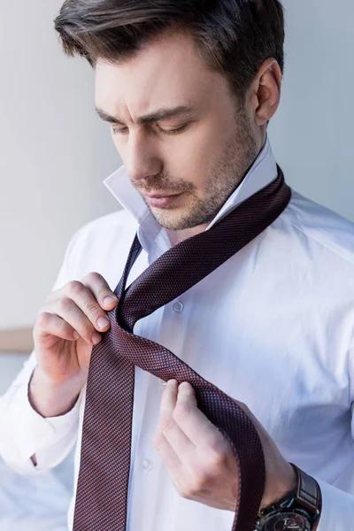 Handsome serious man putting tie on while standing near bed at home — Stock Photo