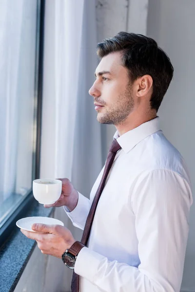 Guapo hombre serio con taza de café de pie junto a la ventana y mirando hacia otro lado - foto de stock