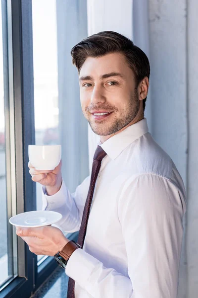 Smiling handsome man standing by window, holding coffee up and looking at camera — Stock Photo