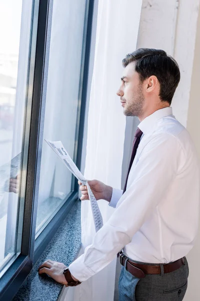 Hombre seguro de buen aspecto de pie junto a la ventana con el periódico y mirando hacia otro lado - foto de stock