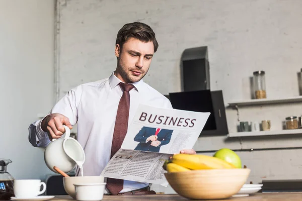 Handsome man reading business newspaper while standing by kitchen table and pouring milk into bowl — Stock Photo