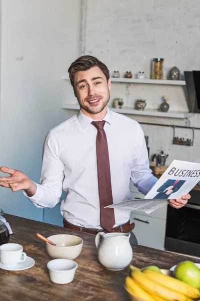 Handsome man standing by kitchen table, holding business newspaper and looking at camera — Stock Photo