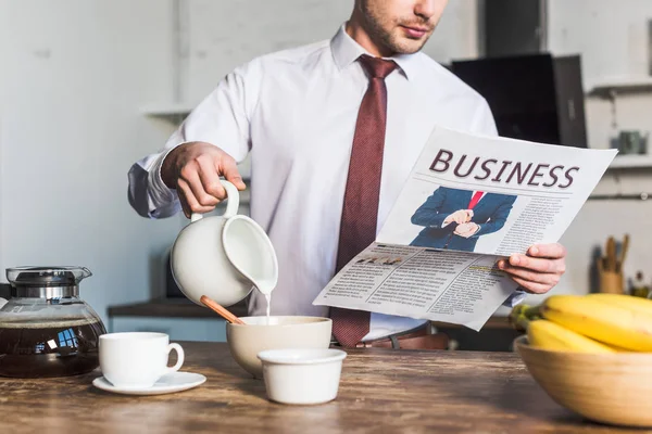 Vue recadrée de l'homme lisant le journal d'affaires tout en se tenant près de la table de cuisine et versant le lait dans le bol — Photo de stock