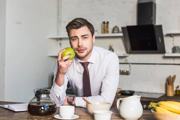 Bell'uomo che tiene la mela e guarda la macchina fotografica mentre è in piedi in cucina a casa — Foto stock