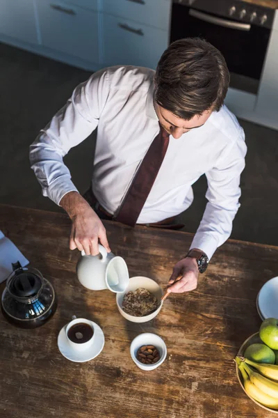 Vista aérea de homem bonito derramando leite em tigela com muesli enquanto toma café da manhã em casa — Fotografia de Stock