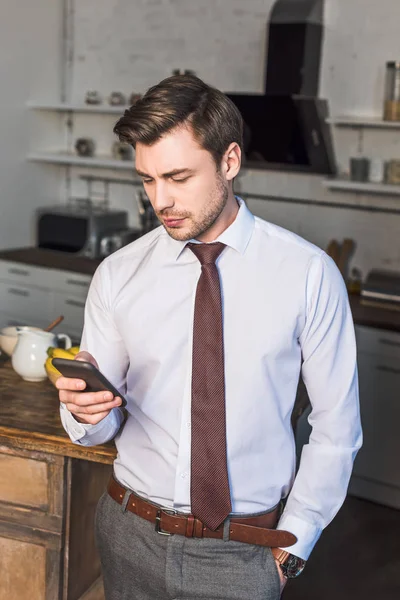 Guapo hombre confiado sosteniendo teléfono inteligente mientras está de pie en la cocina en casa - foto de stock