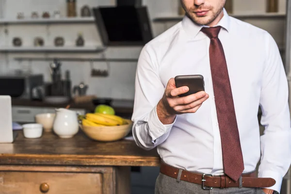 Частичный вид человека с помощью смартфона, стоя на кухне дома — стоковое фото