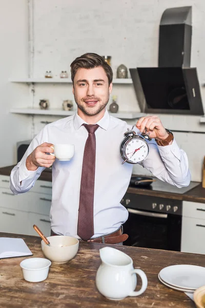 Hombre alegre sosteniendo taza de café y despertador mientras está de pie en la cocina en casa - foto de stock