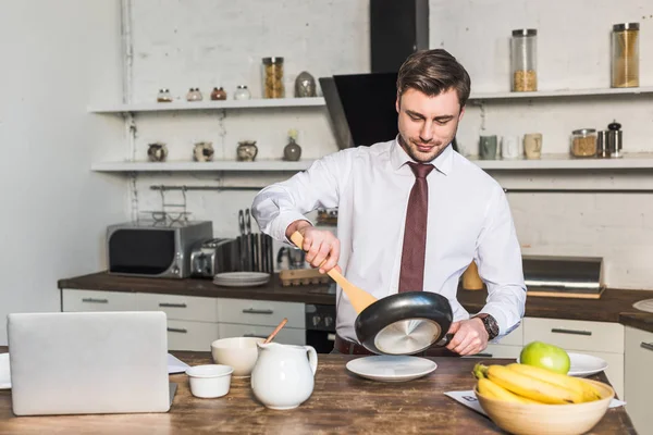Hombre guapo con sartén preparando el desayuno mientras está de pie junto a la mesa de la cocina - foto de stock