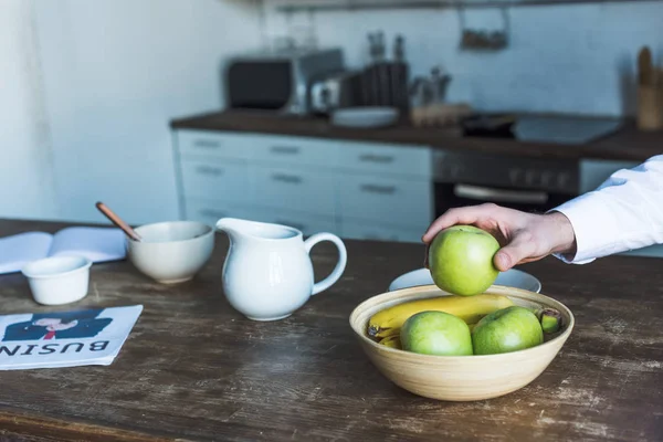 Частичный вид человека, берущего яблоко из миски на деревянном кухонном столе — стоковое фото