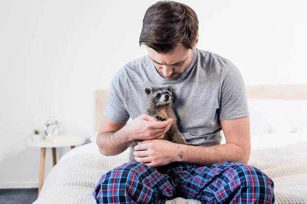 Schöner Mann im Pyjama sitzt auf Bettwäsche und kuschelt mit lustigem Waschbär — Stockfoto