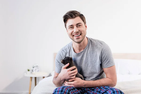 Cheerful man in pyjamas sitting on bedding and cuddling with cute raccoon — Stock Photo