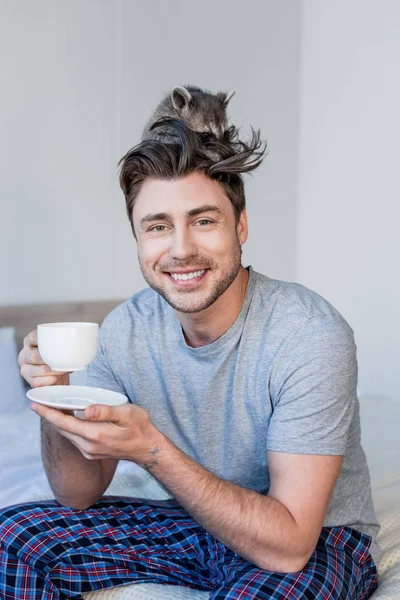Fröhlicher Mann mit lustigem Waschbär auf dem Kopf, Kaffeetasse haltend und in die Kamera blickend — Stockfoto