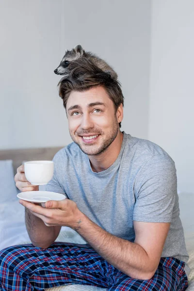 Homme gai avec raton laveur drôle sur la tête tenant tasse de café tout en étant assis sur la literie — Photo de stock