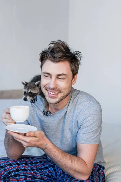 Lächelnder Mann mit lustigem Waschbär auf der Schulter, der Kaffeebecher hält, während er auf Bettwäsche sitzt — Stockfoto