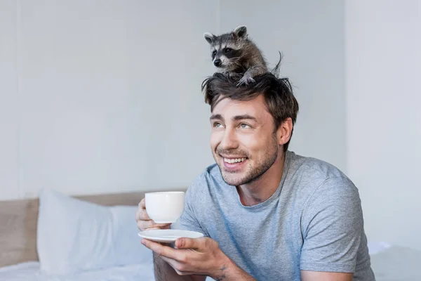 Allegro bell'uomo con procione carino sulla testa che tiene la tazza di caffè mentre si siede su biancheria da letto — Foto stock