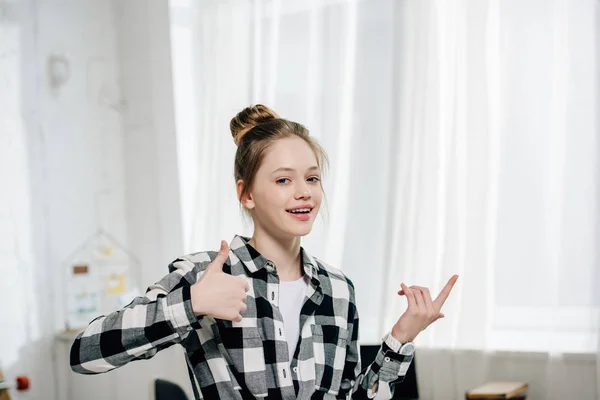 Sonriente adolescente con camisa a cuadros mostrando pulgares en casa - foto de stock