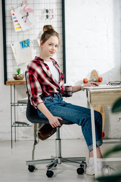 Cute teenage kid in jeans sitting on chair at table and looking at camera — Stock Photo