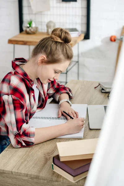 Teenager in checkered shirt writing in notebook while doing homework — Stock Photo