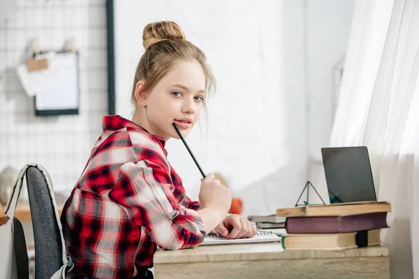 Pensive teenager in checkered shirt holding pen at table while doing homework — Stock Photo