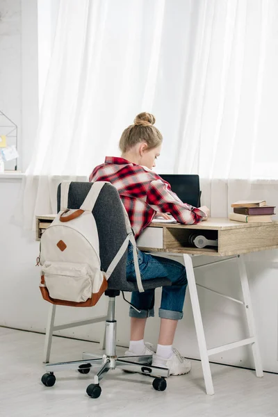 Adolescente con camisa a cuadros sentado en el escritorio y haciendo la tarea - foto de stock