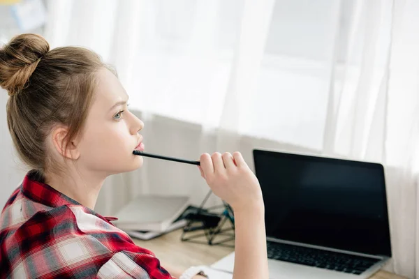 Pensive teenage kid with pen sitting at table with laptop — Stock Photo