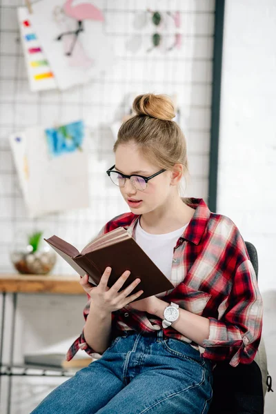 Curious teenage kid in glasses and checkered shirt reading book — Stock Photo