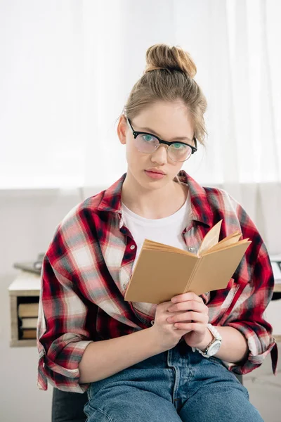 Curioso adolescente con gafas y camisa a cuadros sosteniendo libro y mirando a la cámara - foto de stock