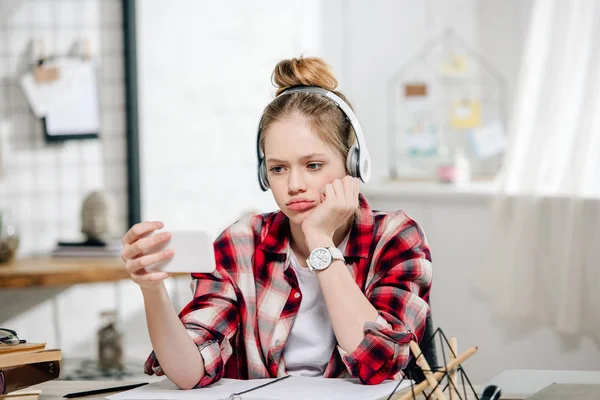 Adolescente triste con camisa a cuadros y auriculares mirando el teléfono inteligente durante la videollamada - foto de stock