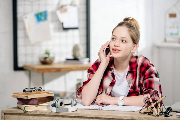 Teenage kid in red checkered shirt sitting at desk and talking on smartphone — Stock Photo