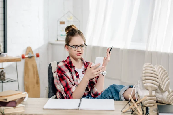 Adolescent dans des lunettes assis avec les jambes sur la table et en utilisant un smartphone — Photo de stock