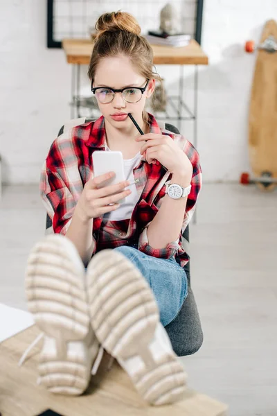 Подросток в очках сидит с ногами на столе и использует смартфон — стоковое фото