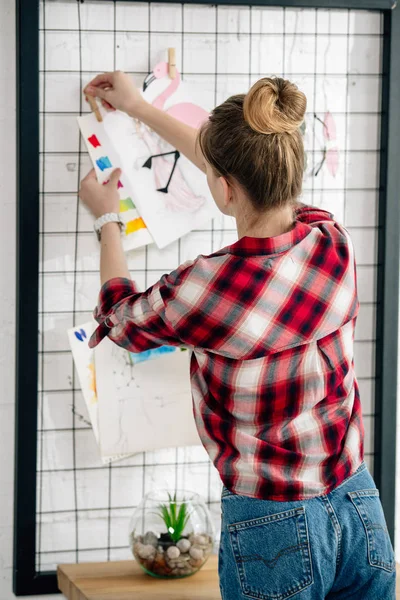 Back view of teenager in checkered shirt hanging drawing on pinboard — Stock Photo