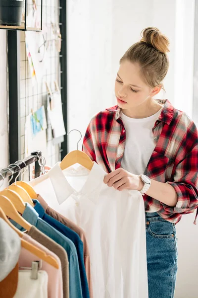 Interested teenage kid in wristwatch holding hanger with white shirt — Stock Photo