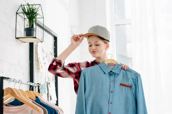 Adolescente niño en gorra sosteniendo percha con camisa azul en casa - foto de stock