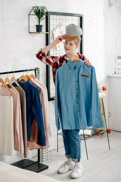 Pleine longueur vue de l'adolescent enfant en chapeau tenant cintre avec chemise bleue à la maison — Photo de stock