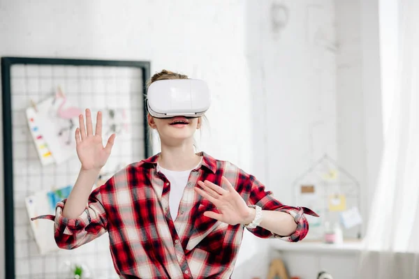 Adolescente con camisa a cuadros roja y auriculares vr gestos en casa - foto de stock