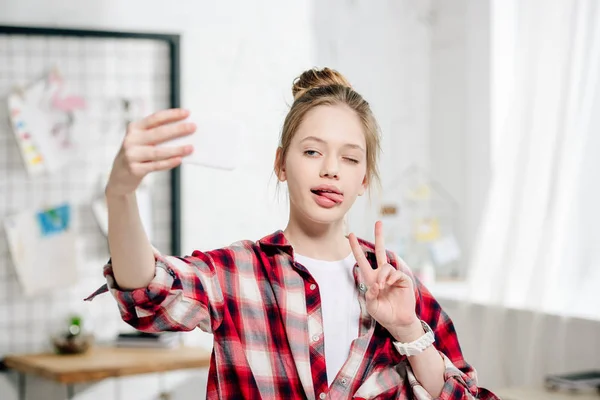 Alegre adolescente niño sosteniendo teléfono inteligente y tomando selfie con signo de paz en casa - foto de stock