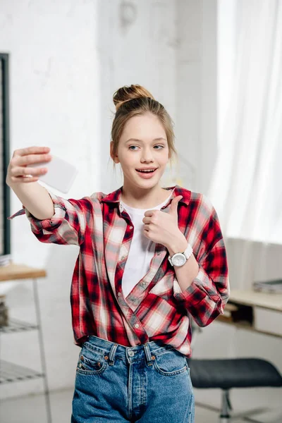 Cheerful teenager kid holding smartphone and taking selfie with thumb up at home — Stock Photo