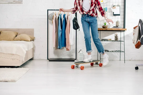Cropped view of teenager in jeans and checkered shirt standing on longboard in bedroom — Stock Photo
