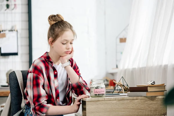 Trauriges Teenie-Kind sitzt am Tisch und blickt auf Sparschwein — Stockfoto