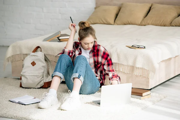 Подросток с ноутбуком и книгами сидит на ковре и делает домашнее задание — стоковое фото