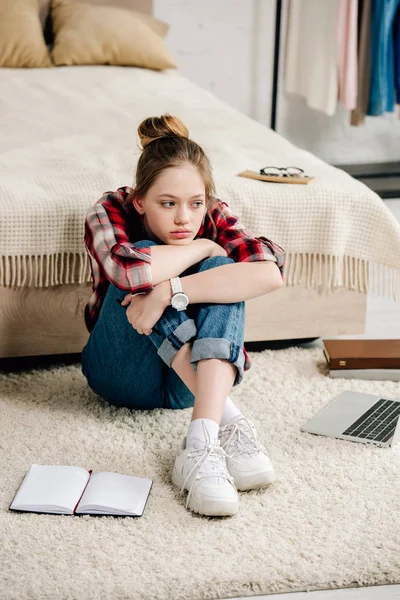 Adolescente ennuyée en chemise à carreaux assise près du lit et embrassant les genoux — Photo de stock