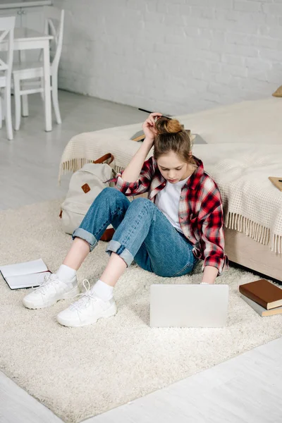Adolescente com laptop e livros sentados no tapete e fazendo lição de casa — Fotografia de Stock