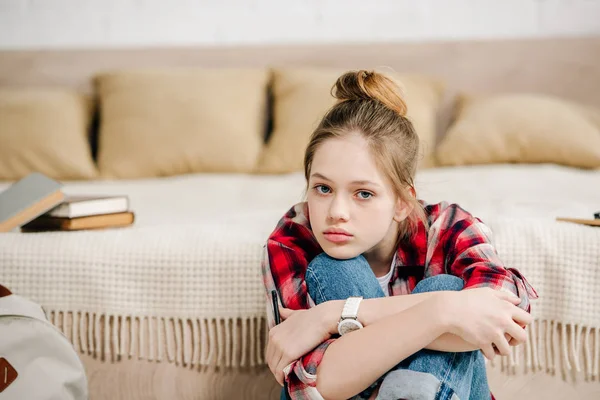 Adolescente ennuyée en chemise à carreaux assise près du lit et embrassant les genoux — Photo de stock